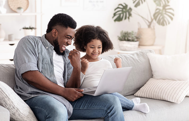 Un padre y su hija están sentados juntos en el sofá, visiblemente emocionados por algo que ven en la computadora portátil que comparten.