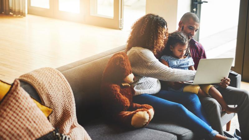 Dos padres y su hija miran una computadora portátil mientras están sentados juntos en un sofá