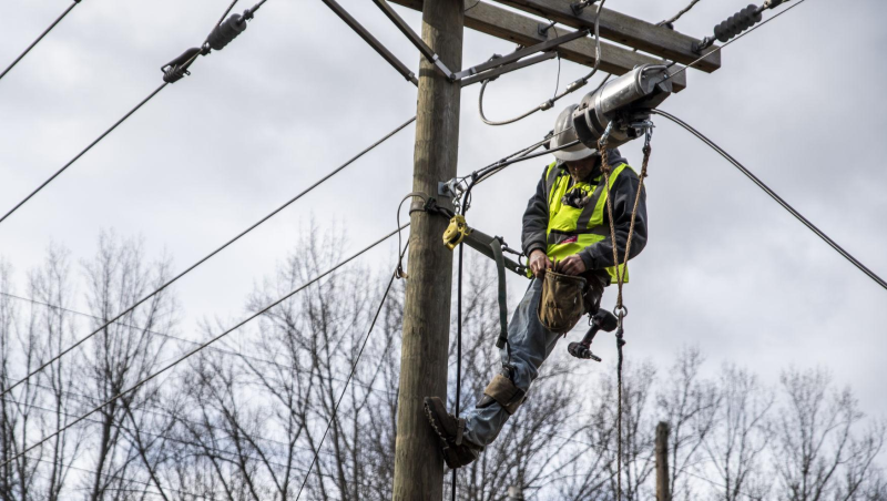 Un trabajador de línea con casco blanco y chaleco de alta visibilidad de color amarillo brillante está sujeto de forma segura a una línea eléctrica y trabaja en ella.